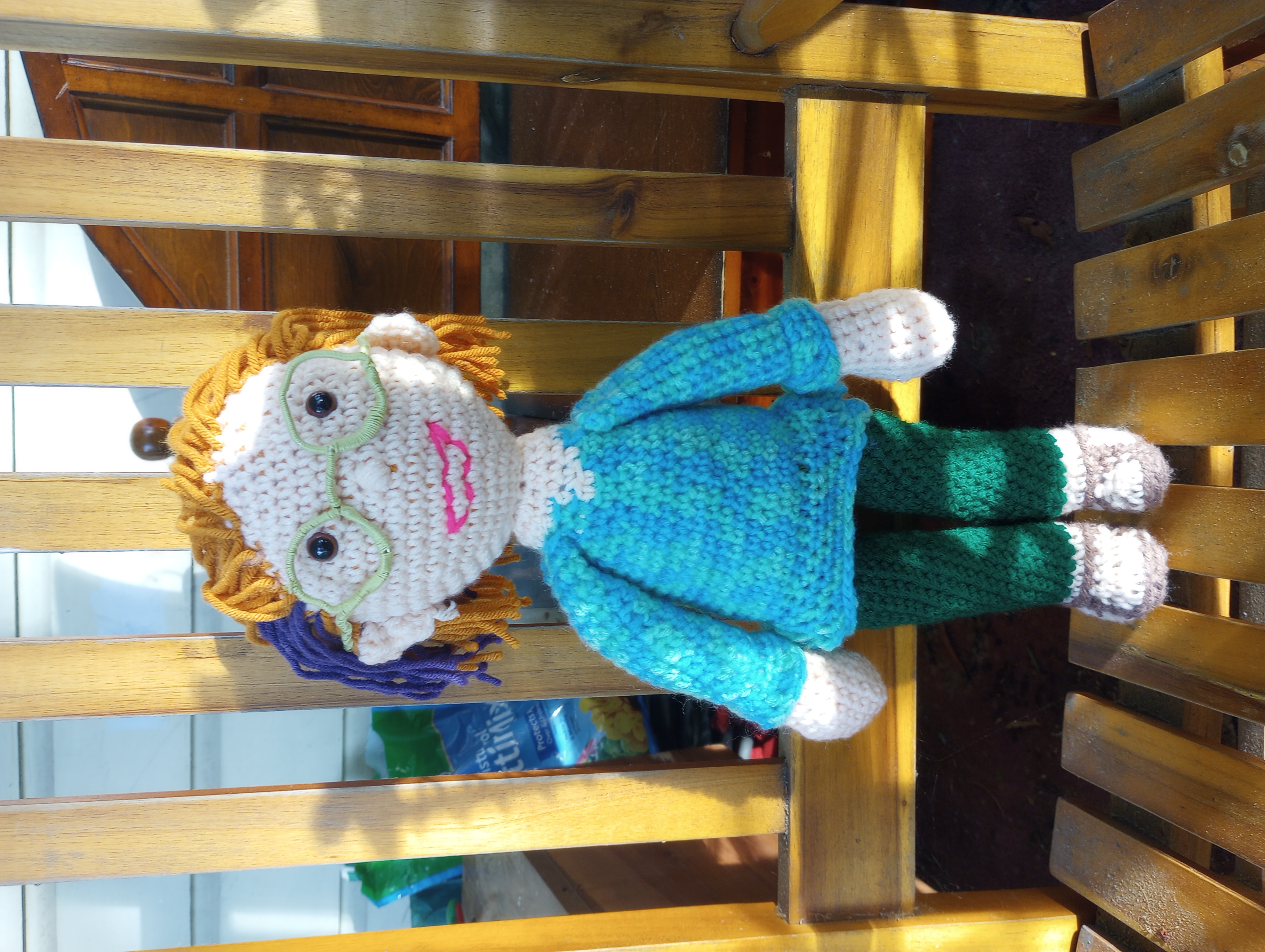 A crocheted doll with peach skin, brown eyes, and orange hair with a purple streak that is tucked behind the ears, wearing a turquoise and green mottled tunic, emerald green leggings, brown sandals, and round pale green glasses, with a neon pink grin