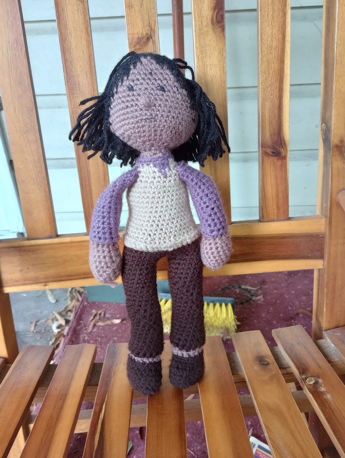 A crocheted doll with light brown skin, long black hair, black eyes, a tan vest, a purple shirt, brown pants, and brown shoes. His pant legs do not meet the top of his shoes.