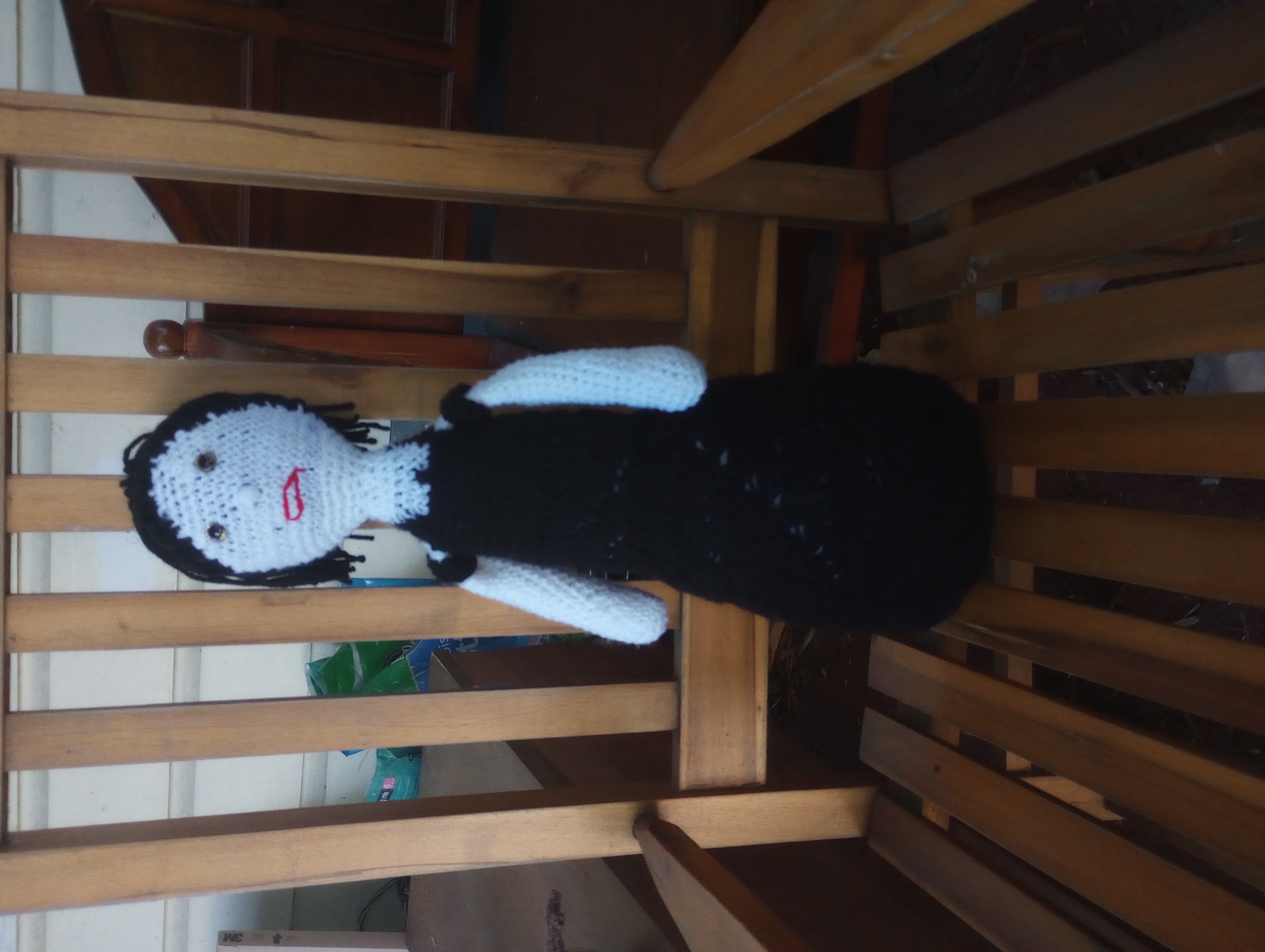 A crocheted doll with white skin and a black off-the-shoulder dress that goes all the way to the floor, with black hair pulled up in a high ponytail, gold eyes, and red lips.