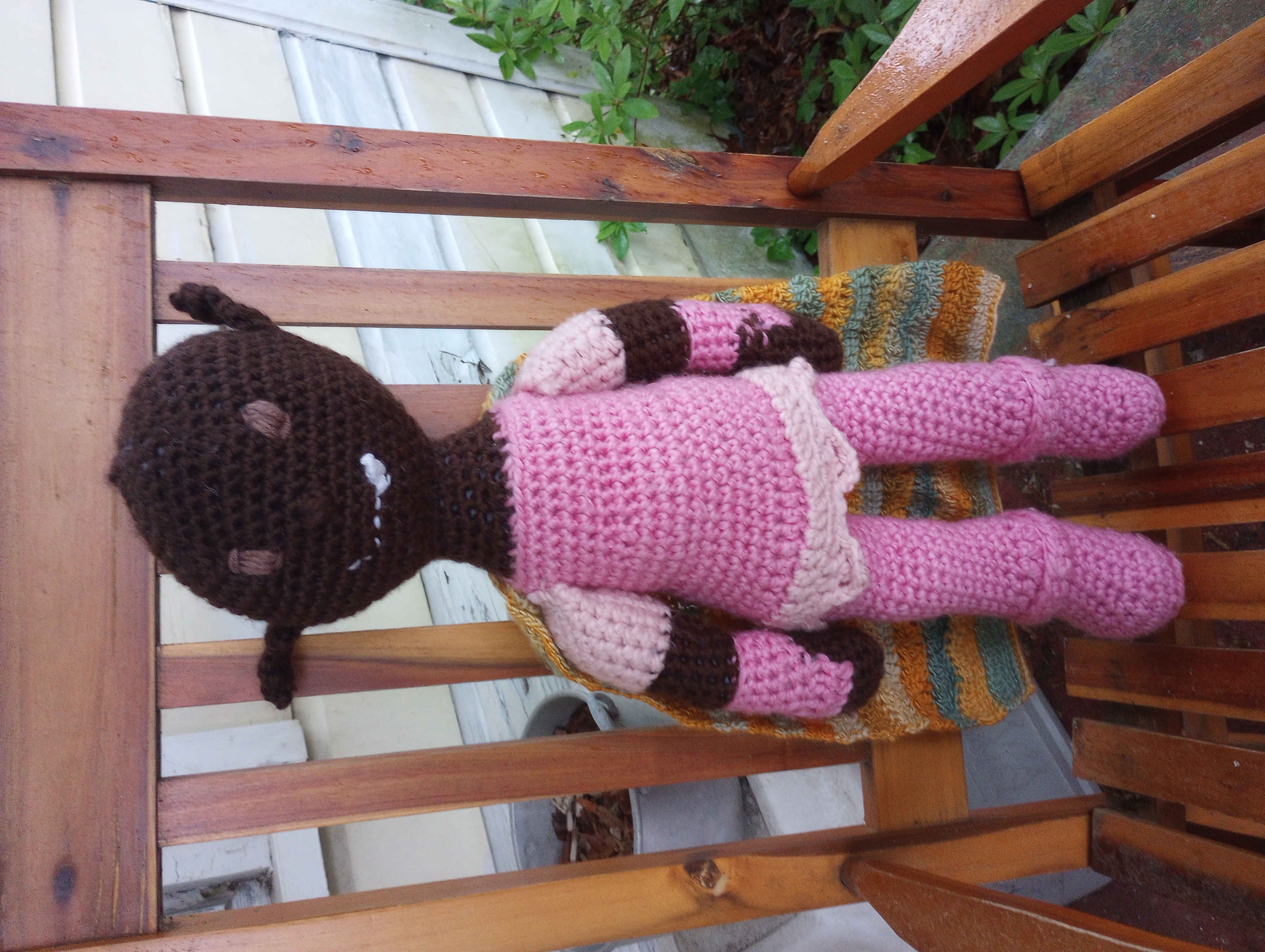 A crocheted doll with dark brown skin, pointed ears, a smile with a single pointed fang, light brown eyes, pink armor with lighter pink sleeves and trim around the waistline, and a gold-green multicolored cloak hanging down the back