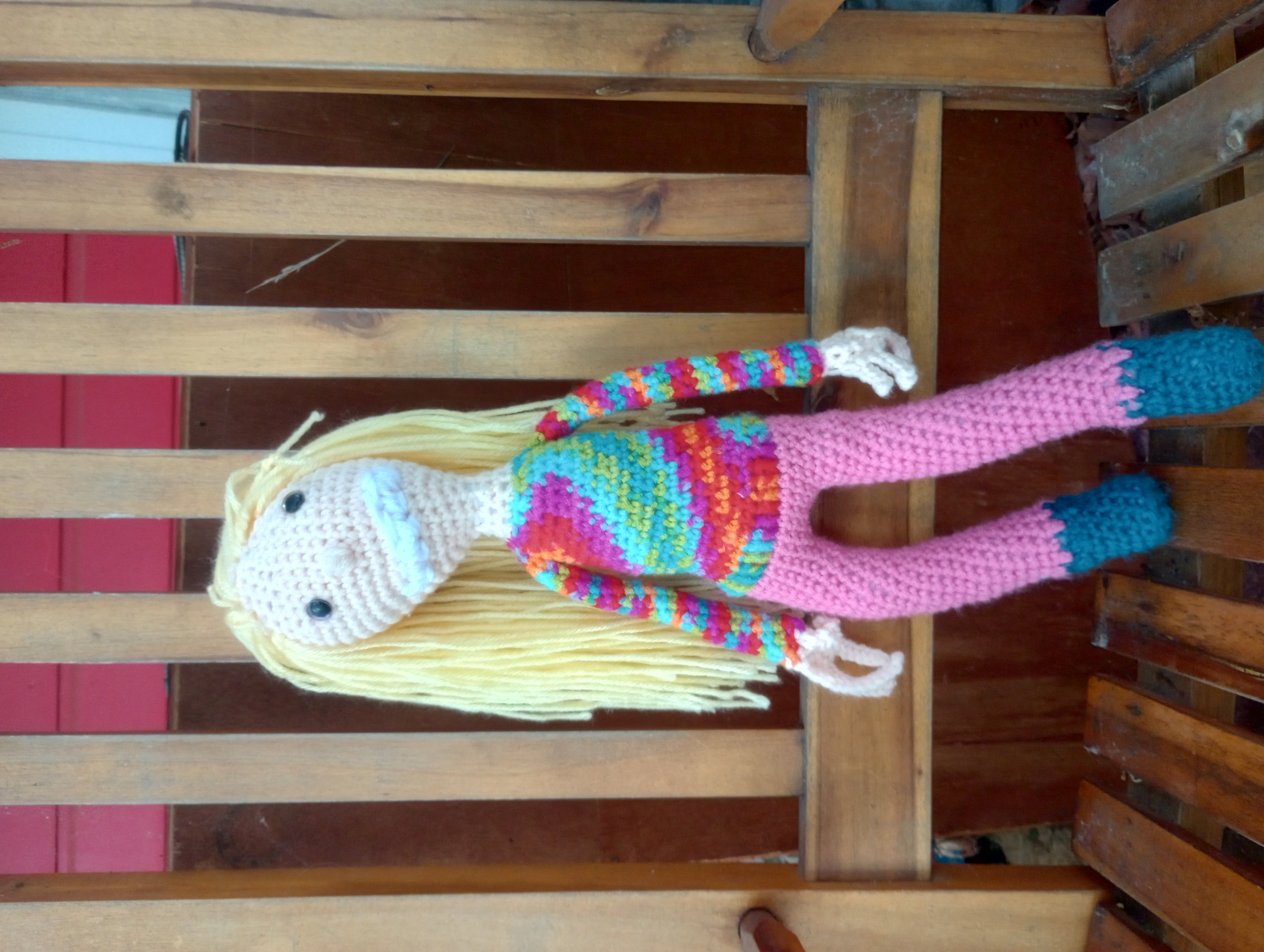 A crocheted doll of Michael the Distortion from The Magnus Archives, a human-shaped being with long yellow hair and pale skin, one purple eye and one gold one, a long thin face, and long skinny arms, wearing teal shoes, pink pants, and a rainbow spiral-colored shirt. It is grinning with far too large a smile for its face.