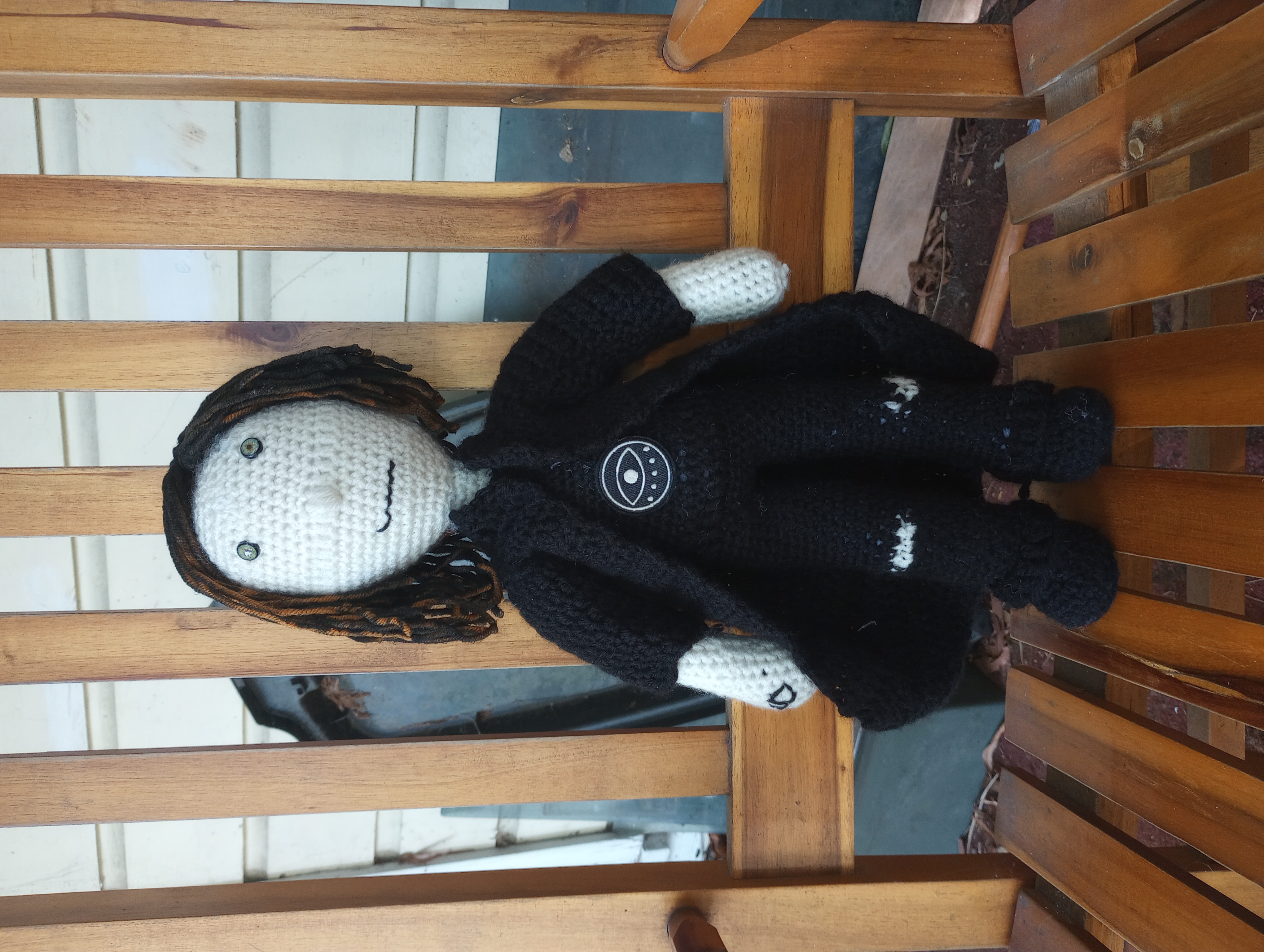 A crocheted doll with a white face and hands, black shirt, pants, and trench coat, hazel eyes, hair made of orange yarn spray-dyed black, and a patch of an eye on the shirt