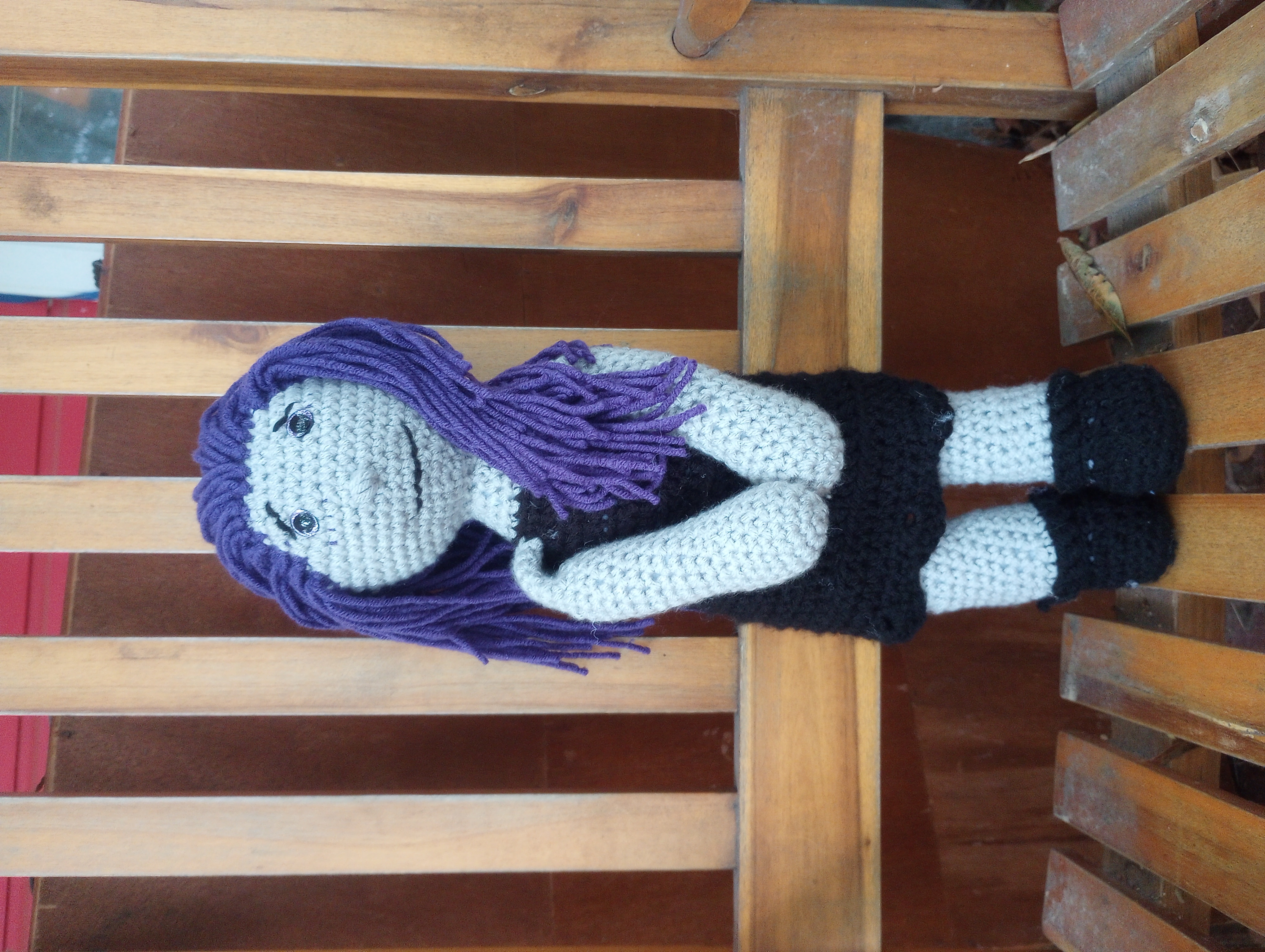A crocheted doll of Isadora Ravenwood, a half Dark Elf with blue-grey skin and purple hair and eyes, wearing a sleeveless black dress and black boots, clasping her hands and looking anxiously at the camera