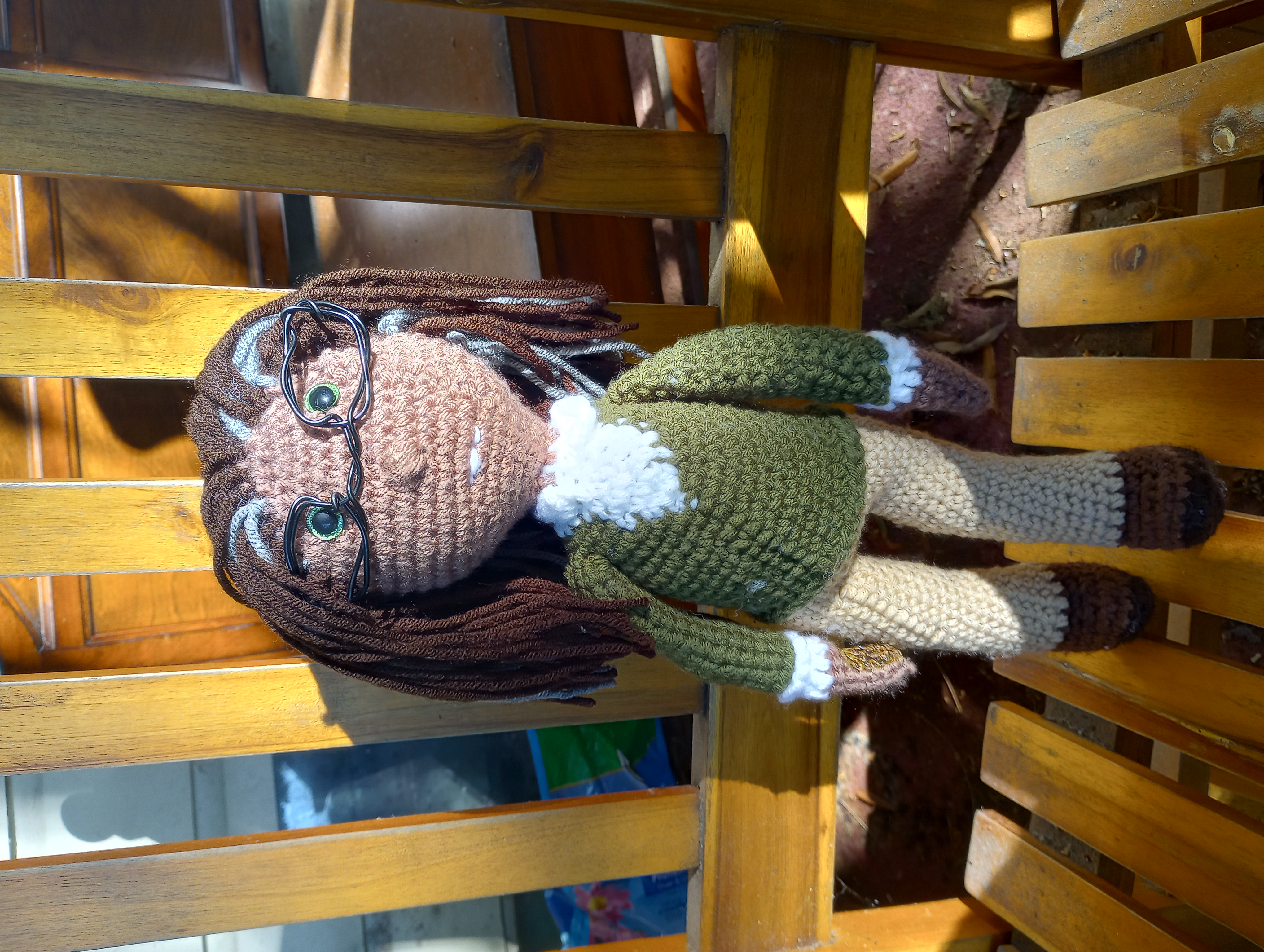 A crocheted doll with light brown skin, dark brown and grey hair, and green eyes, wearing a white collared shirt, a green cardigan, khaki trousers, brown shoes, and black square glasses