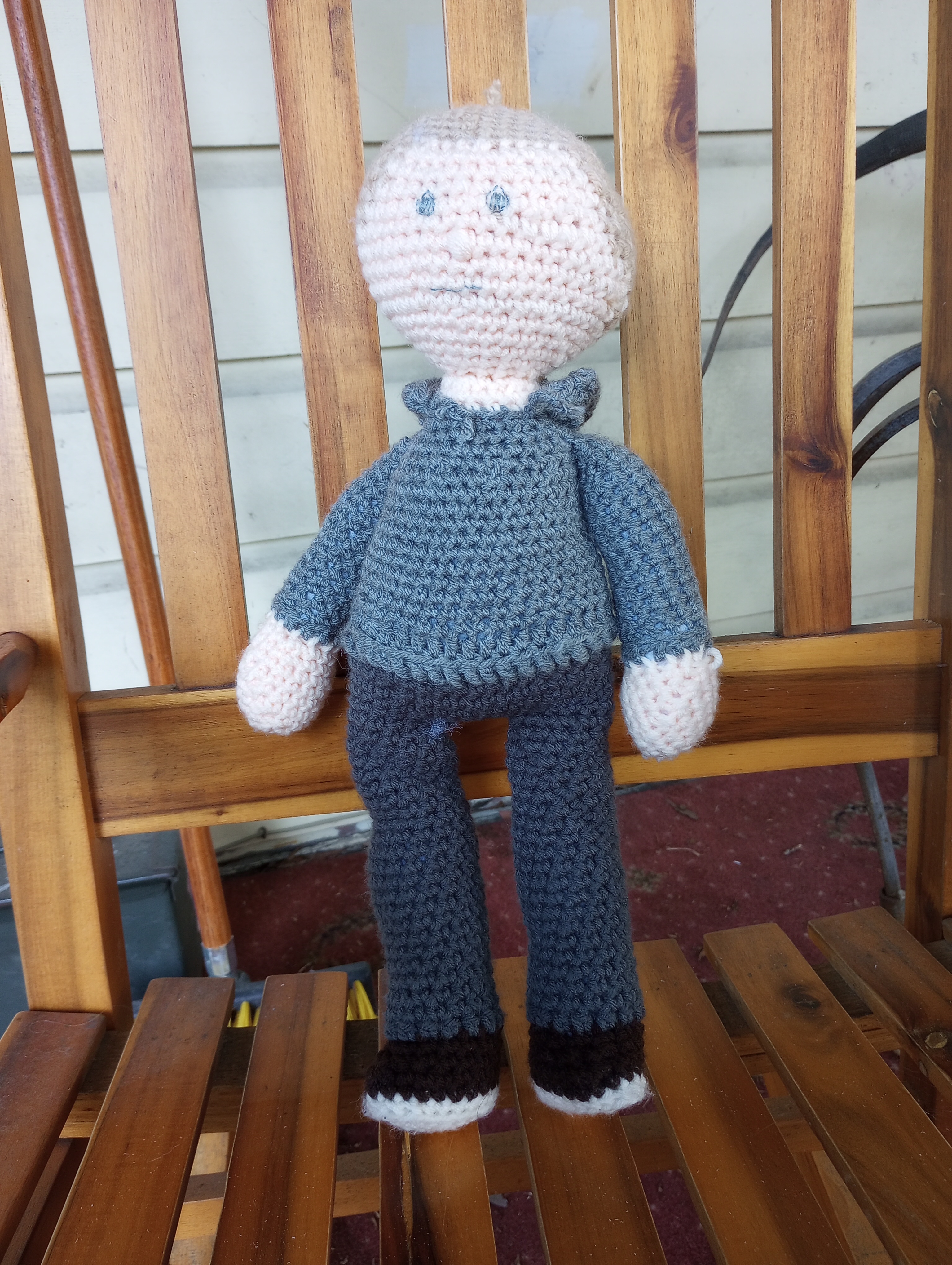 A crocheted doll with pale skin, pale blond hair flush with the scalp, a dark grey hoodie, grey sweatpants, dark brown sneakers, grey eyes, and a straight grey mouth