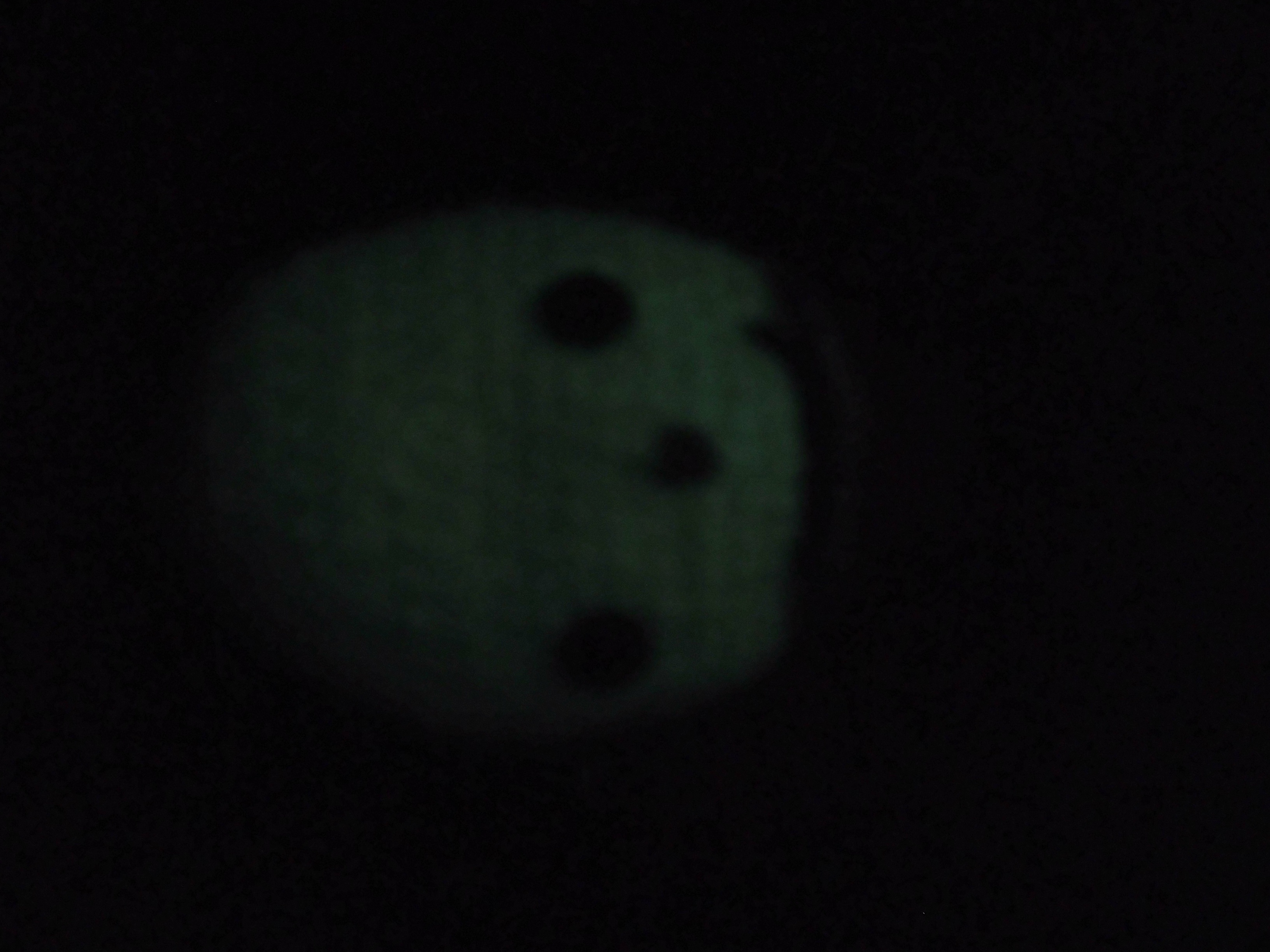 A close up of the doll's head in the dark. It is glowing an eerie green, except for dark holes where the eyes, nose, and mouth are