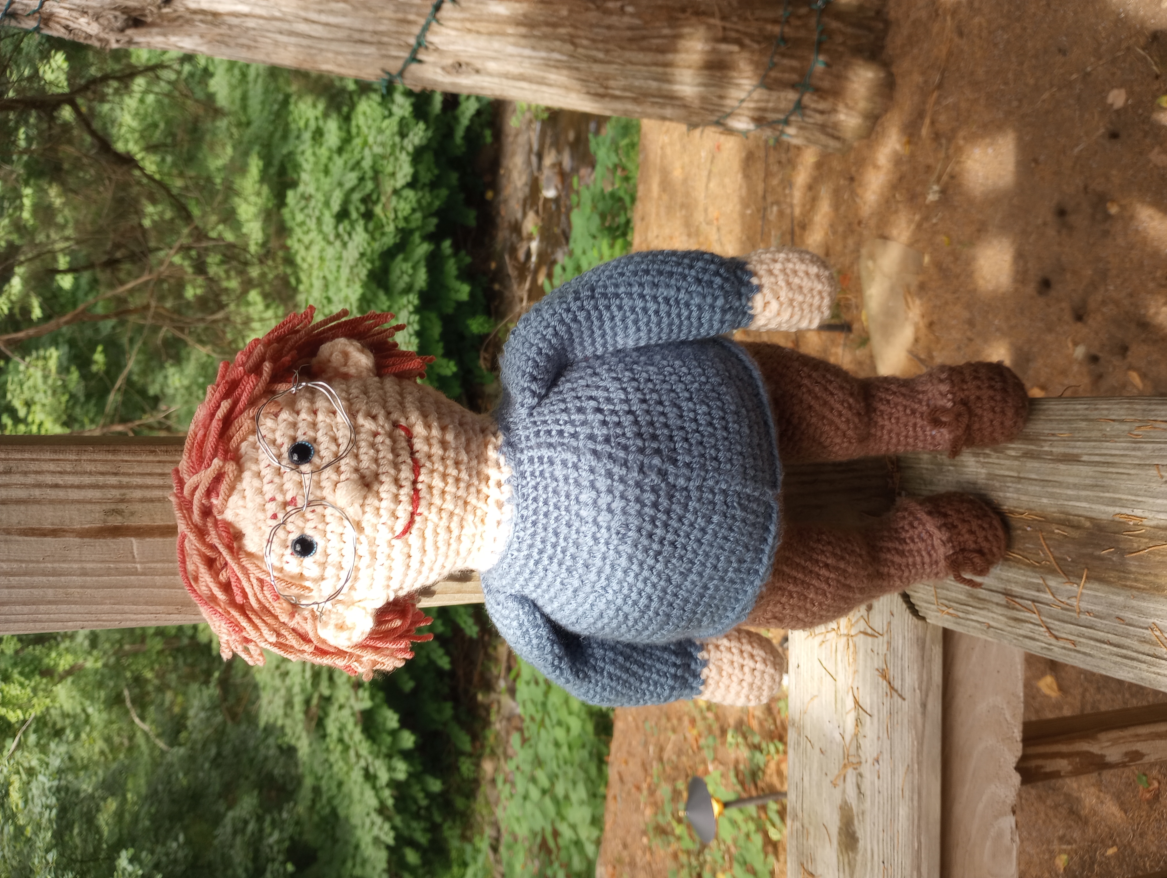 A crocheted doll with pale skin, blue eyes, and ginger hair gradually seeming to go grey, wearing brown trousers, a blue jumper, and silver glasses.