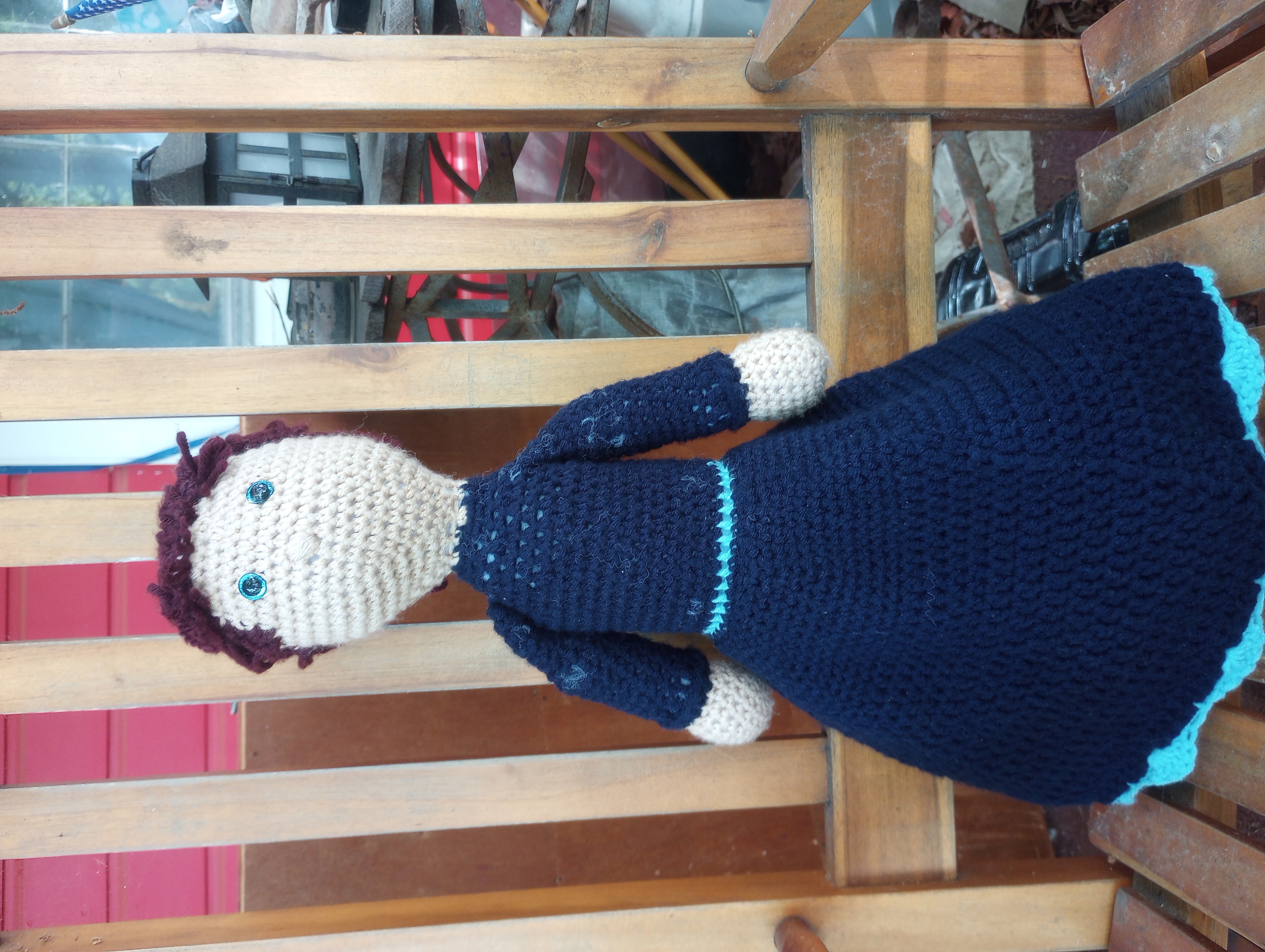 A crocheted doll with slightly tanned skin, glittery blue eyes, reddish-brown hair pulled back in an elegant hairstyle, and a navy blue high-necked dress with a turquoise sash and ruffle at the hem