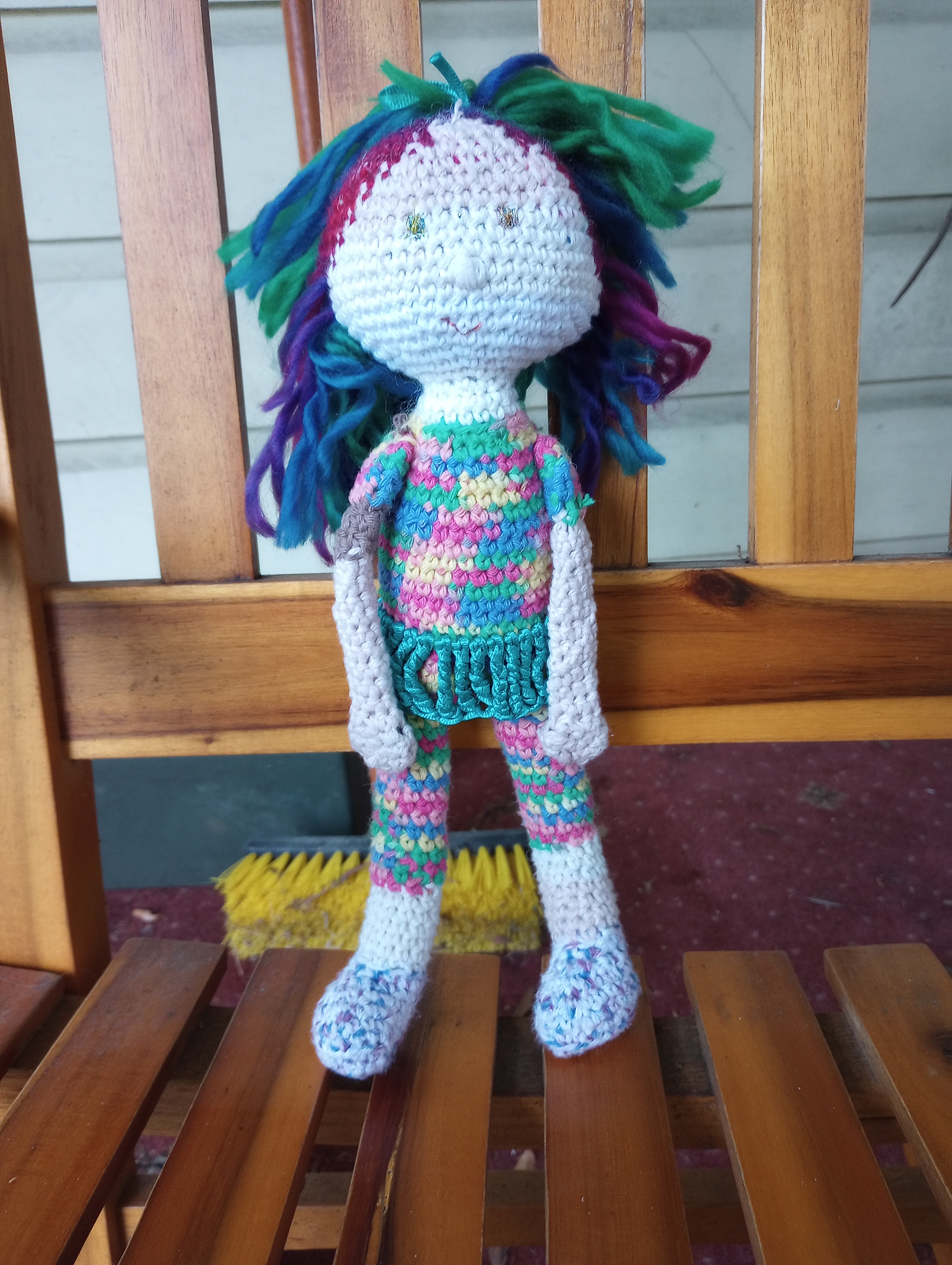 A crocheted doll with pale skin in variegated cream stripe yarn, sparkly eyes, long fluffy rainbow-colored hair pulled into a half-ponytail on top, blue and white spotted shoes, and a rainbow-colored leotard with a teal ribbon skirt.