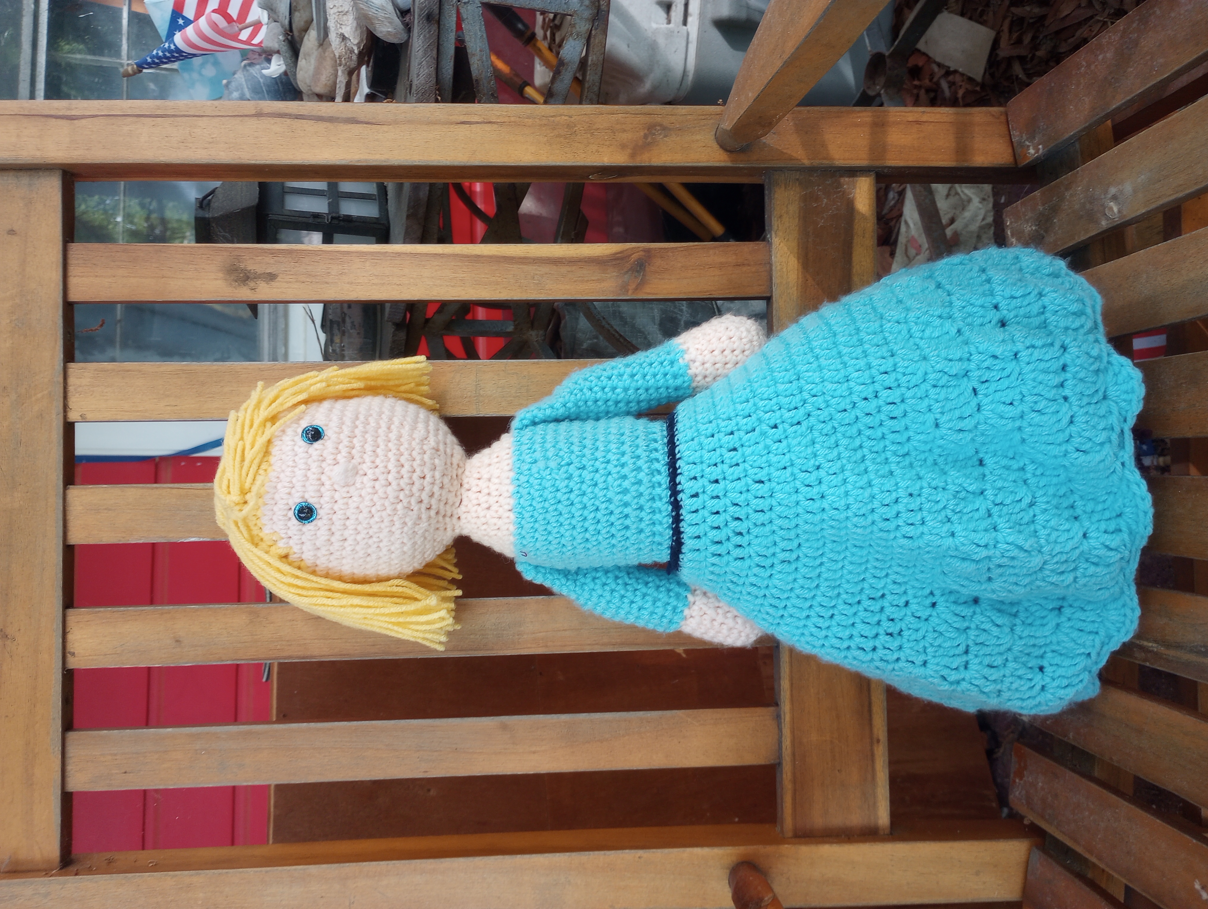 A crocheted doll with pale white skin, glittery blue eyes, yellow hair parted on one side that comes down to her chin, and a turquoise skirt and blouse with a navy blue belt/sash at the waistline