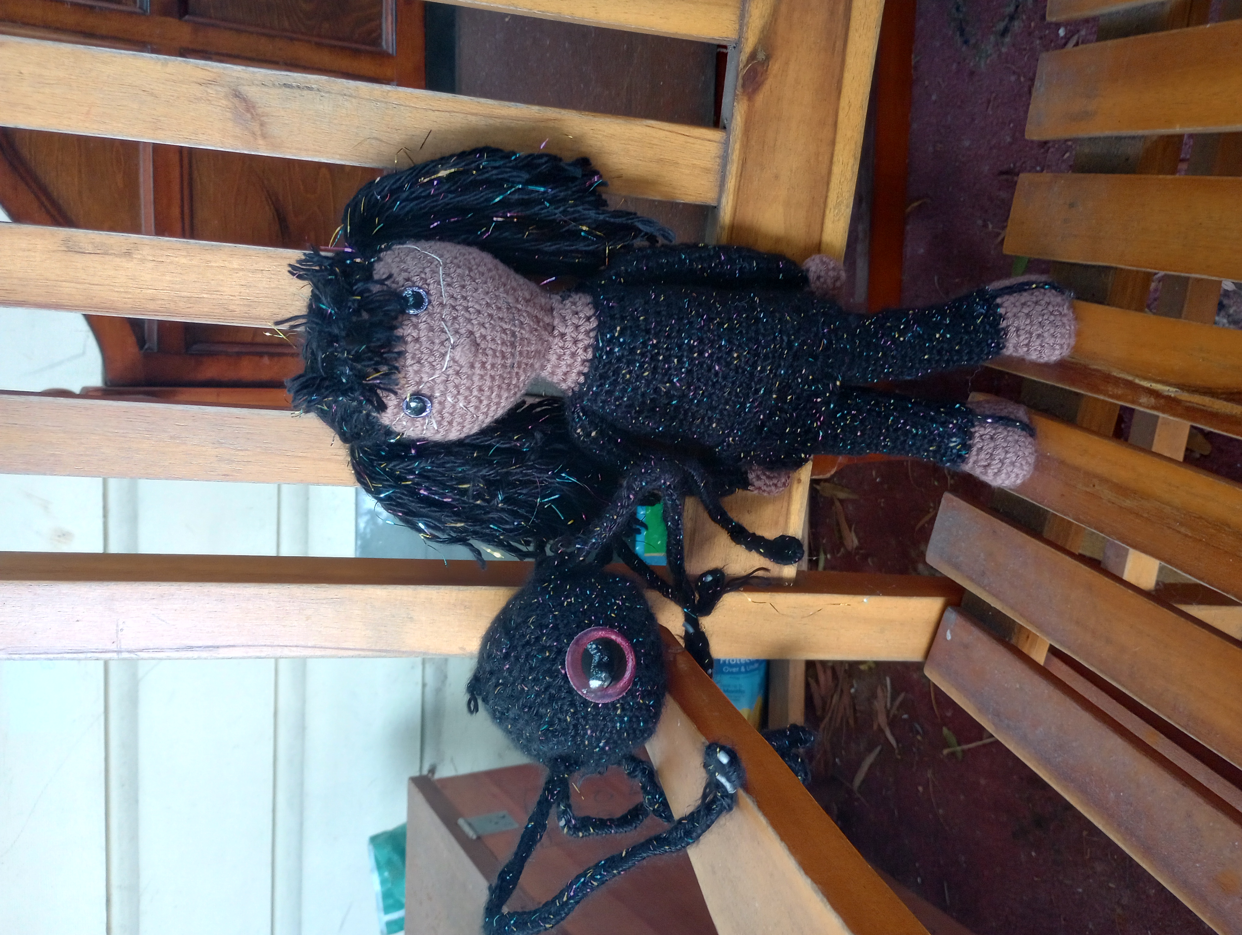 A crocheted doll with tanned skin, black pigtails with rainbow filaments running through them, and black clothes that match her hair. She is standing next to a crocheted robot of the same yarn, rectangular with six spiderlike legs and a single red eye