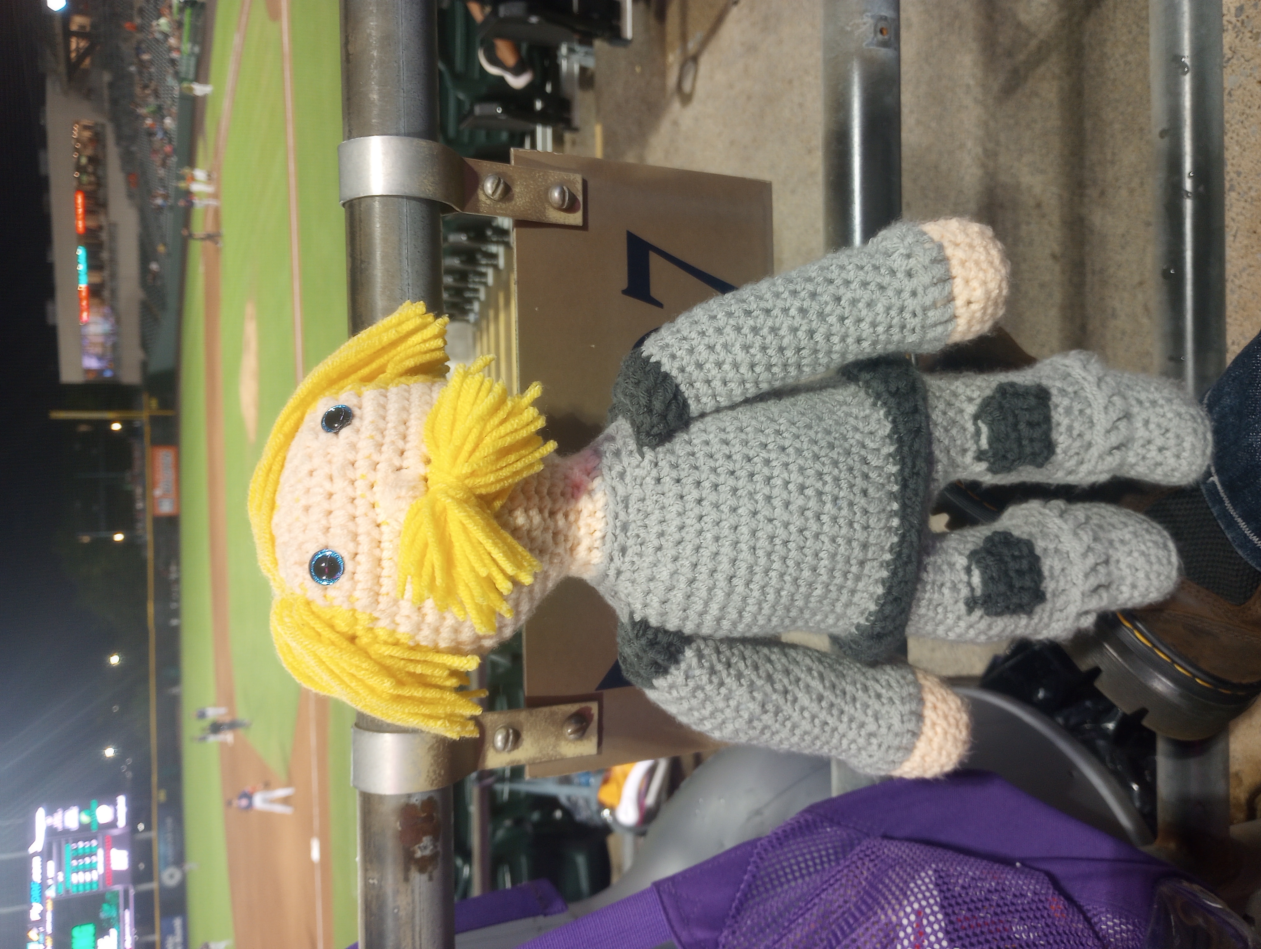 A crocheted doll of a fantasy Dwarf with light skin, bright blue eyes, blond hair cut in a heroic bowl cut, and a bushy blond mustache, wearing grey armor with dark grey pauldrons, kneepads, and trim around the breastplate