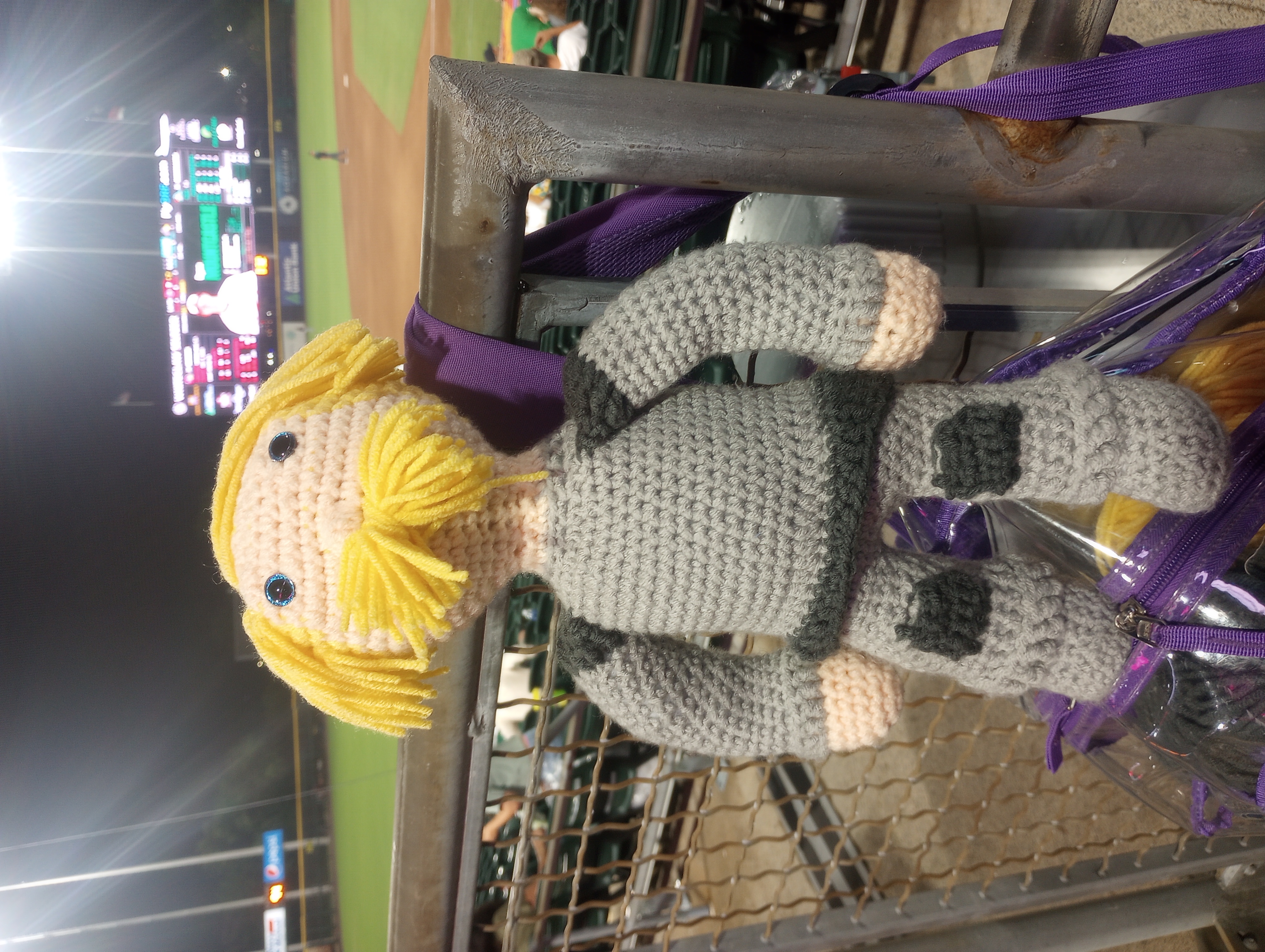 The same doll, standing triumphantly with his arms more or less propped on his hips atop a purple backpack hanging off a railing