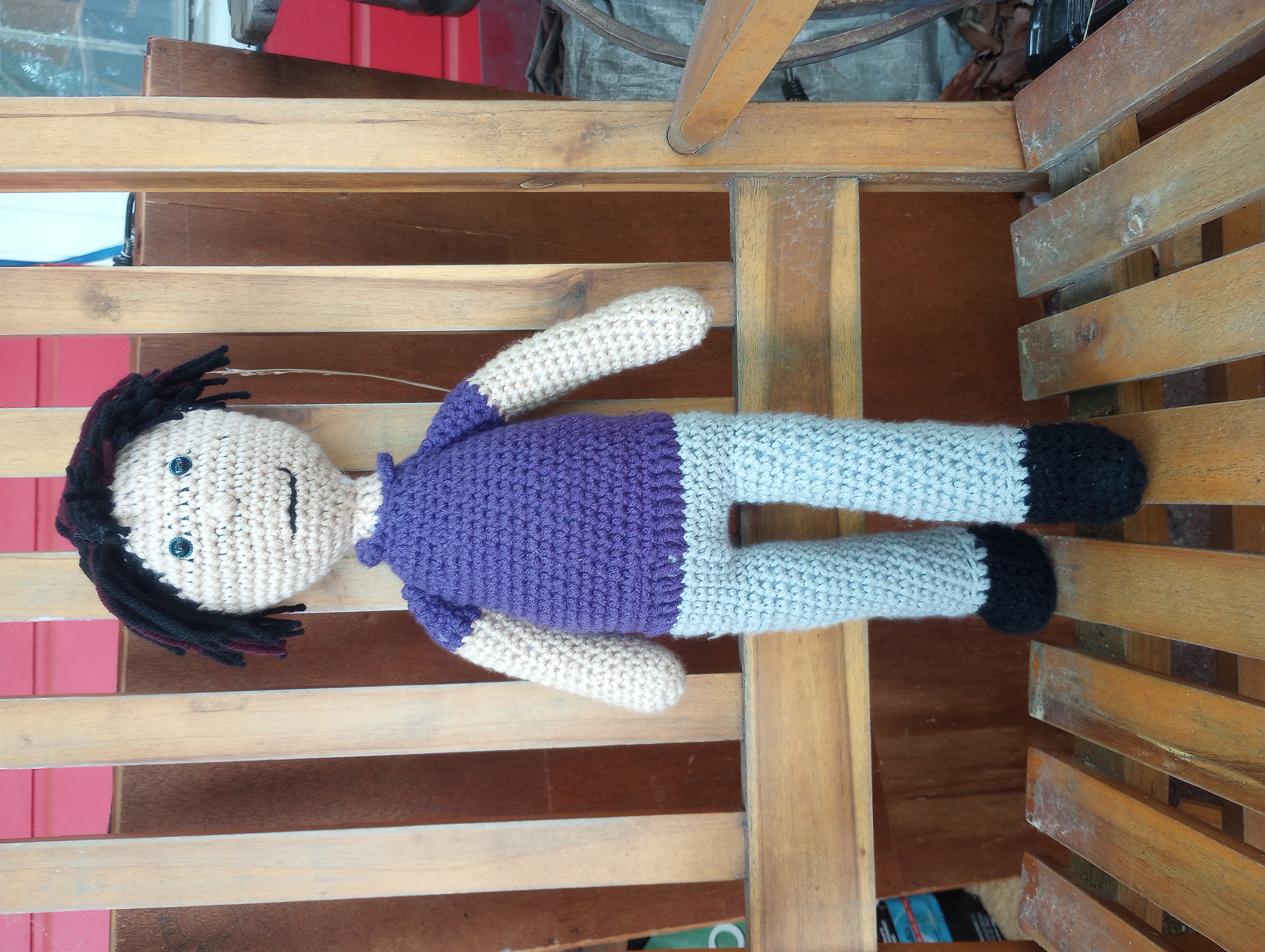 A crocheted doll with lightly tanned skin, black hair streaked with auburn, and blue eyes, wearing a purple short-sleeved polo shirt, grey trousers, and black shoes