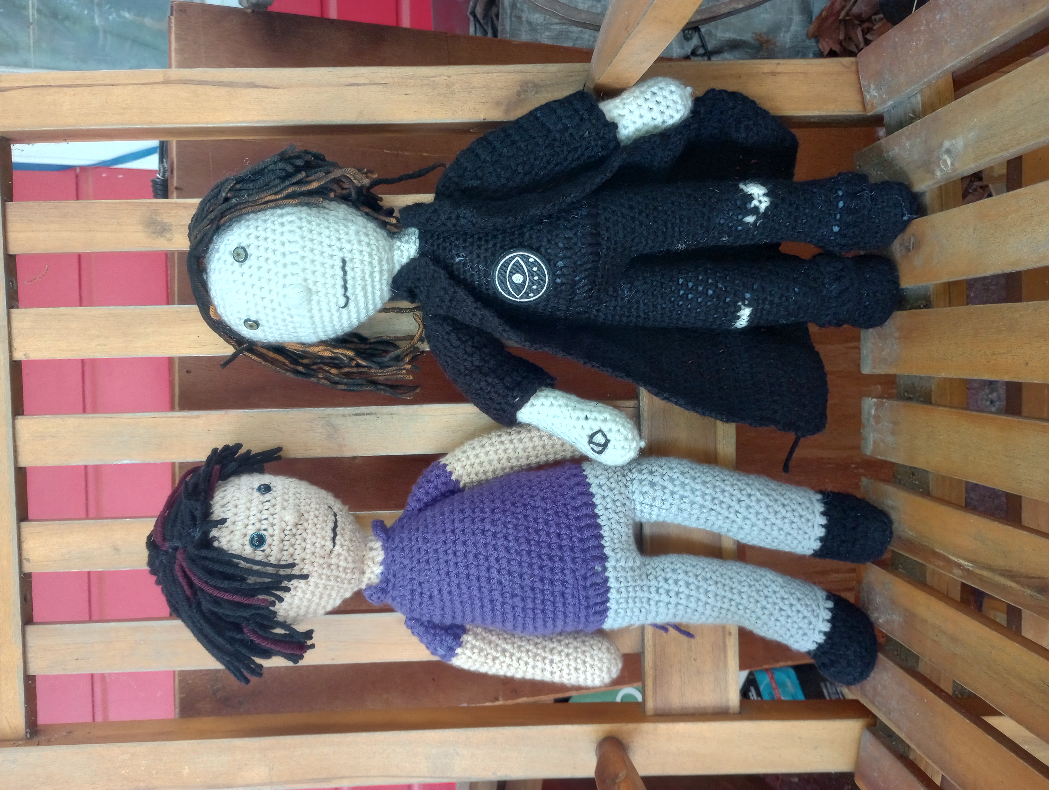 The crocheted doll holding hands with a second doll with pale skin, hazel eyes, and badly dyed black hair with the carrot orange underneath visible, wearing a black t-shirt with an eye on the front, black torn baggy pants, black boots, and a black trench coat